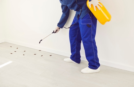 A pest control professional in a blue uniform sprays insects on a light floor against a white wall.