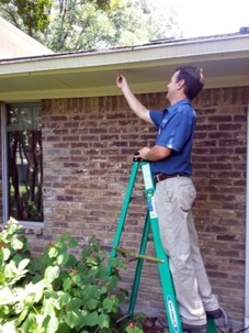 A pest control technician on a ladder inspects the eaves of a brick home for pest activity.