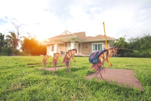 A line of five cockroaches, the last carrying a backpack, marches across a sunny lawn towards a house.